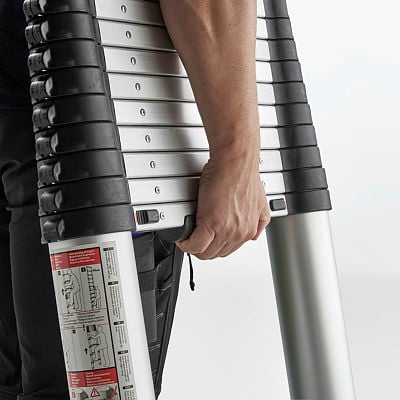 A close-up of a person carrying a telescopic ladder with a compact, collapsible design. The ladder features silver aluminum rungs with black plastic edges and a locking mechanism. The person grips the side of the ladder, showcasing its portability. Safety instructions are visible on the side of the ladder.
