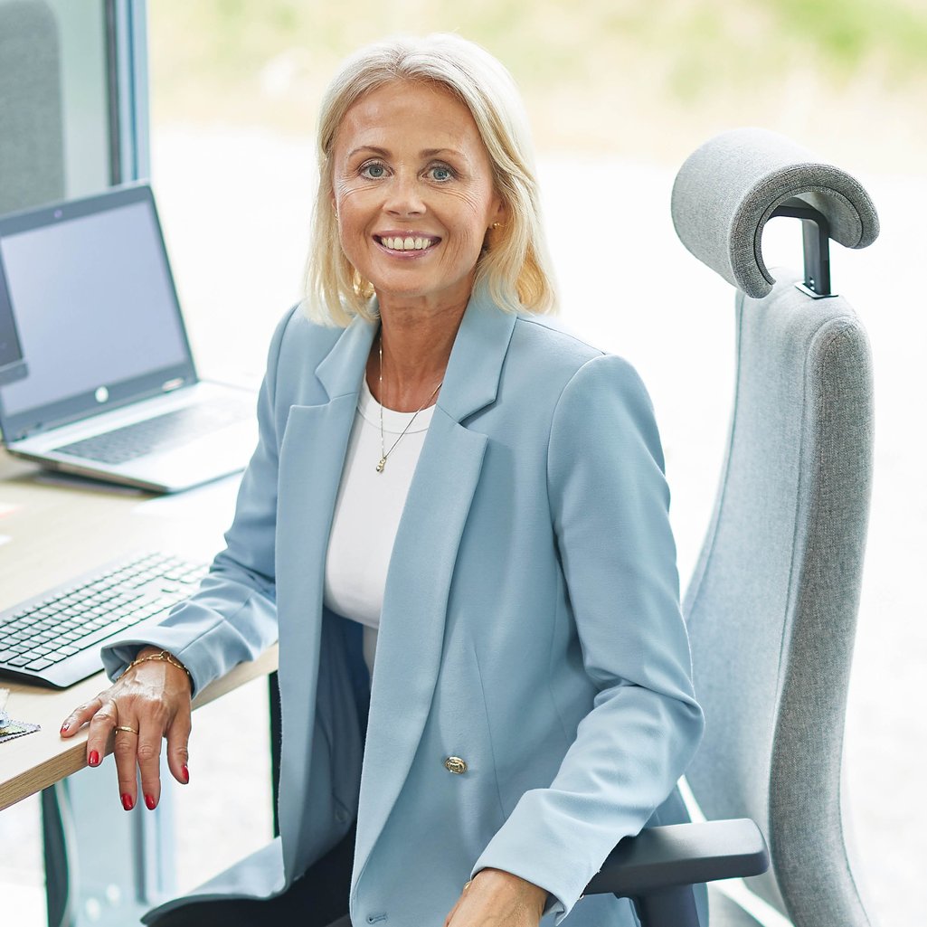 Ženska se smeji v kamero. Ženska ima blond lase in nosi svetlo modro jakno čez belo majico. Na sliki je z dvignjenim pasom, sedi na sivem pisarniškem stolu in se naslanja na mizo s prenosnikom in tipkovnico.