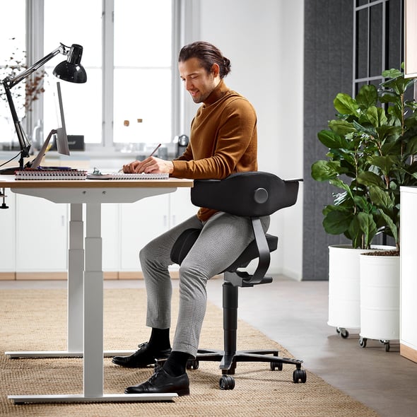 A contemporary workspace featuring a man seated on an ergonomic saddle chair with back support, working at a height-adjustable desk. The setup includes a desk lamp, notebooks, and a laptop, with a bright window and lush green plants enhancing the airy, modern office atmosphere.