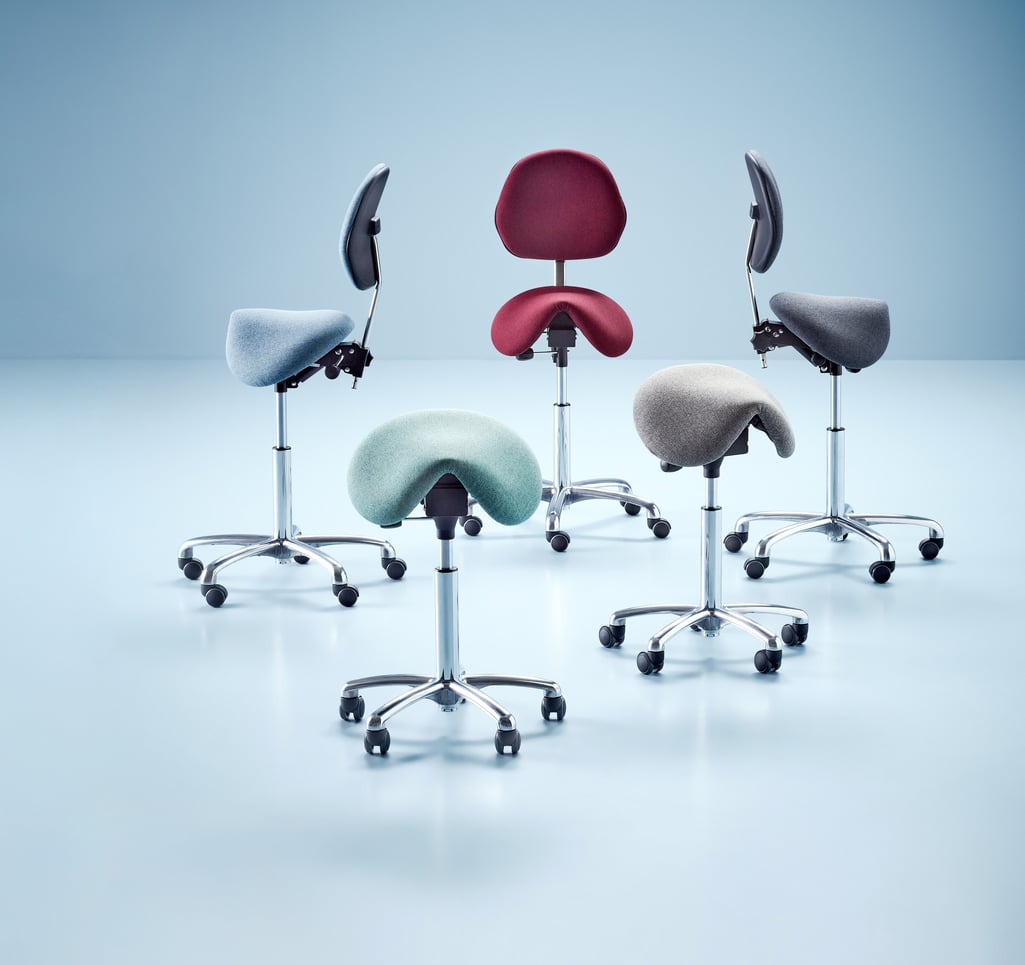 A collection of ergonomic saddle chairs in various colours, including red, grey, blue, and mint green. The chairs feature chrome bases with castors and adjustable heights, with some models including backrests for added support. Set against a clean, light blue background, this arrangement highlights both style and functionality.