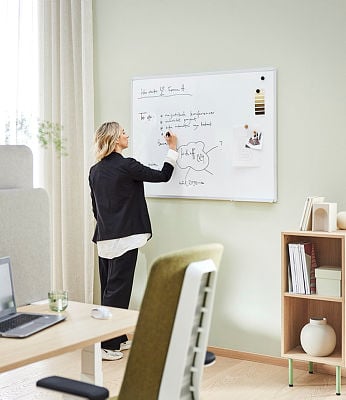 A professional woman with blonde hair, dressed in a black blazer, white blouse, and black trousers, stands in a modern office, writing on a wall-mounted whiteboard. The whiteboard contains handwritten notes, diagrams, and pinned materials, including fabric swatches and a small image. The office is well-lit with natural light coming through a window with sheer curtains. A desk in the foreground has a laptop, a glass of water, and a pair of earbuds. A green office chair with a white frame is partially visible. To the right, a wooden bookshelf holds books, decorative objects, and storage boxes. The overall setting is minimalist and stylish, with soft green walls and light wooden flooring.