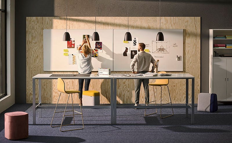 A modern collaborative workspace with two professionals, a woman and a man, working on large wall-mounted whiteboards attached to a plywood-paneled wall. The woman, wearing a light-colored blouse and jeans, is pinning images and notes, while the man, dressed in a button-up shirt and trousers, is drawing a diagram. The space features a high table with a sleek metal frame, yellow bar stools, and various office supplies. Hanging black pendant lights provide focused lighting. The room has a contemporary industrial aesthetic, with a dark carpet, natural wood textures, and a large window allowing natural light to enter. In the background, a white storage cabinet with shelves filled with colorful folders and books is visible, along with poufs and stools for flexible seating.