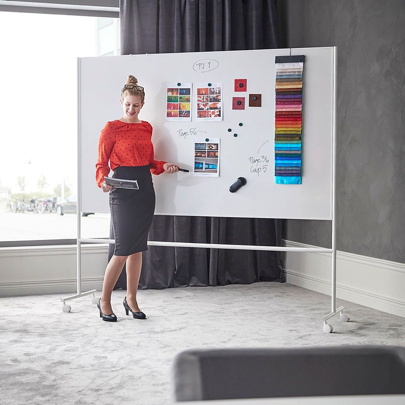 A professional woman in a red polka-dot blouse and black pencil skirt stands in front of a large, wheeled whiteboard in a modern office setting. She holds a black marker in one hand and a folder in the other while presenting. The whiteboard displays images, fabric samples in a range of colors, handwritten notes, and magnets holding up various materials. The room has a grey carpet, a dark curtain, and a window in the background letting in natural light. The woman is wearing black high heels and has her hair styled in a neat updo