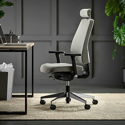 An ergonomic grey office chair with adjustable armrests and a headrest, placed on a textured beige rug in a stylish home office setting. The background features dark panelled walls and lush green plants for a modern and professional aesthetic.