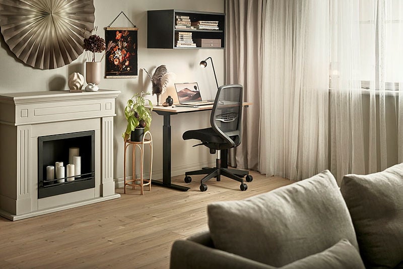 A cozy home office setup featuring a minimalist desk with a black ergonomic chair and a laptop, complemented by a decorative cream fireplace adorned with candles and modern accents. The space includes a beige sofa, soft curtains that filter natural light, and artistic wall decor for a warm and inviting ambiance.