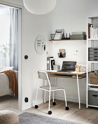 A compact and modern workspace featuring a sleek white desk with a wooden top, paired with a minimalist chair on castors. The setup includes a laptop, desk lamp, and organisational accessories, with shelves above for additional storage. The area is brightened by natural light flowing from an adjacent room, creating a clean and functional atmosphere.