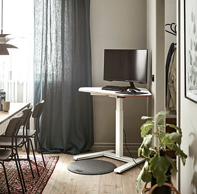 A compact home office setup featuring a height-adjustable white desk with a monitor, keyboard, and mouse. The desk is placed near a window with floor-length dark curtains, complemented by a wooden floor and a cosy rug. A nearby dining area and greenery add a warm and functional touch to the space.