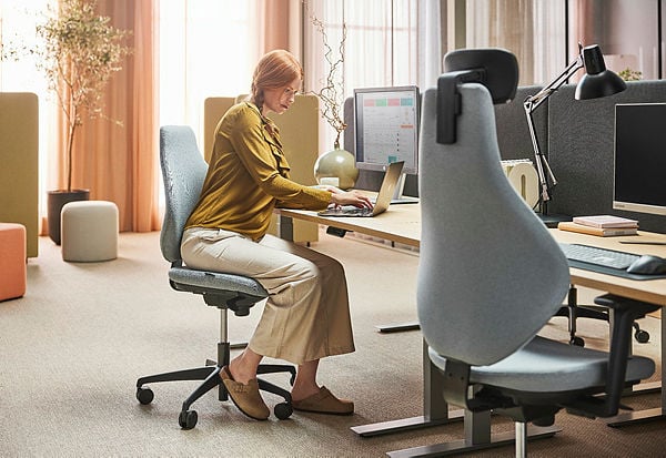 The image depicts a modern office setting with a woman seated at a workstation, working on a laptop. She is wearing a mustard-coloured blouse and beige trousers, seated on an ergonomic office chair with a grey fabric finish. The desk is spacious and organised, featuring a desktop monitor, a task lamp, and a few neatly arranged items. The background includes soft pastel tones with pouffes and tall plants, creating a warm and inviting atmosphere. Natural light streams through sheer curtains, enhancing the comfortable and productive vibe of the workspace.