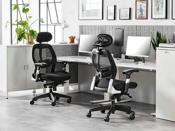 A modern office workspace featuring two black ergonomic mesh chairs with adjustable headrests and armrests. The setup includes a clean, white desk with dual monitors, decorative plants, and neatly organised storage cabinets. The backdrop of grey acoustic panels and framed artwork adds a stylish and professional touch