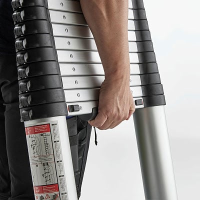 A close-up of a person carrying a telescopic ladder with a compact, collapsible design. The ladder features silver aluminum rungs with black plastic edges and a locking mechanism. The person grips the side of the ladder, showcasing its portability. Safety instructions are visible on the side of the ladder.