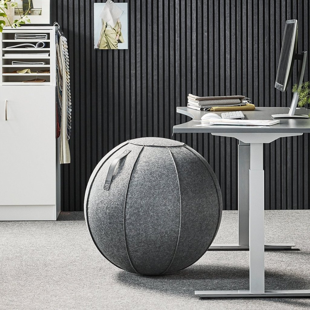 A modern office setup featuring a grey fabric-covered exercise ball chair with a built-in handle, positioned next to a sleek height-adjustable desk. The workspace has a contemporary design with a black slatted wall in the background, adding texture to the interior. A neatly organized white shelving unit holds documents, fabric swatches, and office supplies, while a framed fashion photograph is pinned to the wall. On the desk, a monitor, keyboard, and stacked textiles contribute to the stylish and functional workspace. The overall aesthetic is minimalist and ergonomic, promoting both comfort and movement in the office environment.