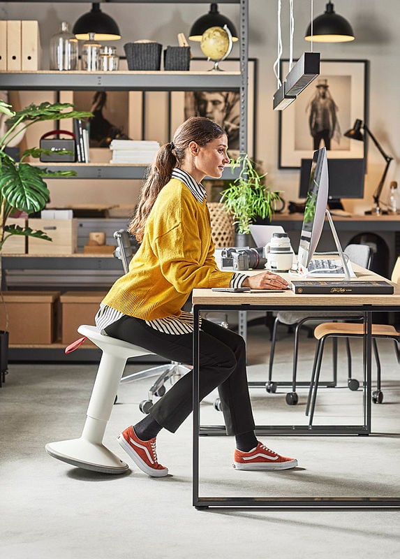 A modern and stylish office space where a woman with long brown hair, wearing a mustard-yellow sweater, striped shirt, black trousers, and red sneakers, is working at a desktop computer. She is seated on a white ergonomic wobble stool, designed for active sitting, leaning slightly forward with a focused expression. The workspace has an industrial aesthetic, featuring a wooden desk with a black metal frame, open shelving with storage boxes, books, and decorative items, and hanging black pendant lights. Large framed black-and-white photographs and green plants add character to the background. The setting combines functionality, comfort, and contemporary design, promoting an ergonomic and creative work environment.