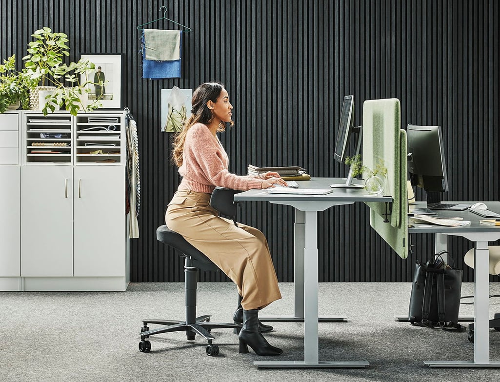 A modern and ergonomic office environment where a woman with long dark hair is working at a height-adjustable desk. She is seated on a black saddle-style ergonomic chair that promotes good posture. Dressed in a pink knitted sweater, beige high-waisted trousers, and black heeled boots, she maintains a focused expression while using a desktop computer. The office features a black slatted wall, a white storage unit with shelves holding fabric samples and office supplies, and a workspace partitioned with green acoustic panels. Small plants and framed artwork add warmth to the contemporary setting, which is designed for both comfort and productivity.