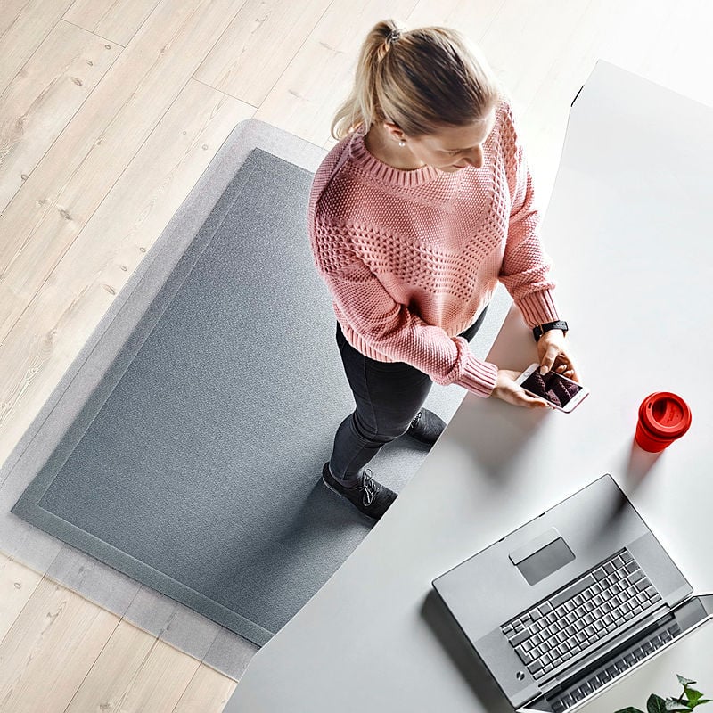 A top-down view of a woman standing at a white desk in a modern workspace. She is wearing a pink knitted sweater, black jeans, and black sneakers. She is holding a smartphone in both hands while looking at the screen. A silver laptop is open on the desk, next to a red reusable coffee cup. She is standing on a grey anti-fatigue mat placed on a light wooden floor, designed to provide comfort for prolonged standing. The setting is minimalistic and contemporary, promoting an ergonomic work environment.