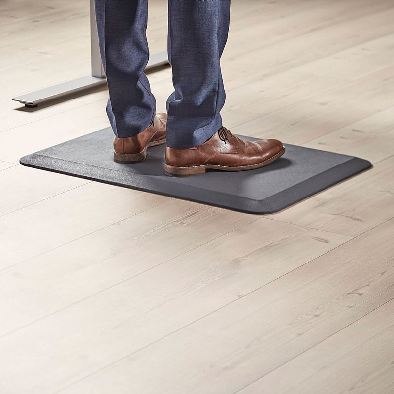 A close-up view of a man’s legs and feet as he stands on a black anti-fatigue mat in an office setting. He is wearing navy blue trousers and brown leather dress shoes. The mat is placed on a light wooden floor and is designed to provide ergonomic support for prolonged standing. Part of a height-adjustable desk leg is visible in the background, suggesting a standing workstation setup. The image highlights workplace comfort and ergonomic solutions.