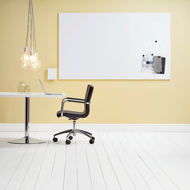 A minimalist office space with a large whiteboard mounted on a soft yellow wall. A few papers and fabric samples are pinned to the board with magnets. Below, a sleek black office chair with chrome detailing is positioned next to a round white table with a chrome base, which holds a laptop and a glass jar. A cluster of exposed filament light bulbs hangs from black cords, adding a warm and modern touch. The floor is made of white wooden planks, enhancing the bright and airy feel of the workspace. The setting is simple yet stylish, promoting a clean and creative work environment.