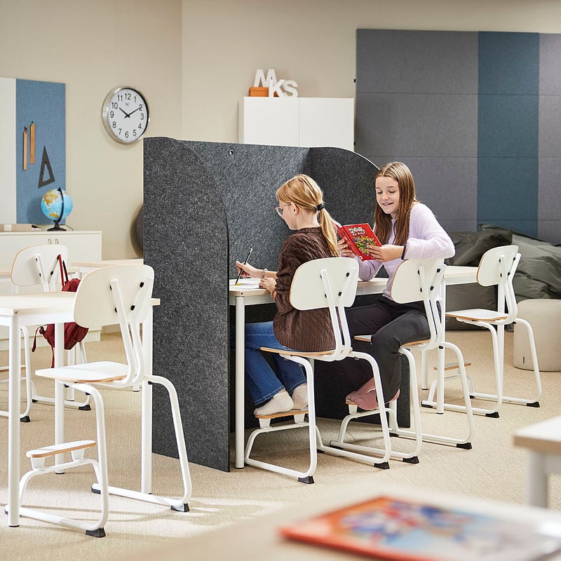 A modern classroom with a flexible learning environment where two students are sitting at a shared desk with a tall, dark grey acoustic divider. One student, wearing a brown sweater and glasses, is focused on writing, while the other, in a light purple sweater, is smiling and holding a colorful book. The classroom is furnished with white chairs and desks, with additional seating and a cozy lounge area with bean bags in the background. A clock on the wall, a globe, and various educational materials create a stimulating yet comfortable atmosphere. The setting promotes both focused work and collaborative learning.