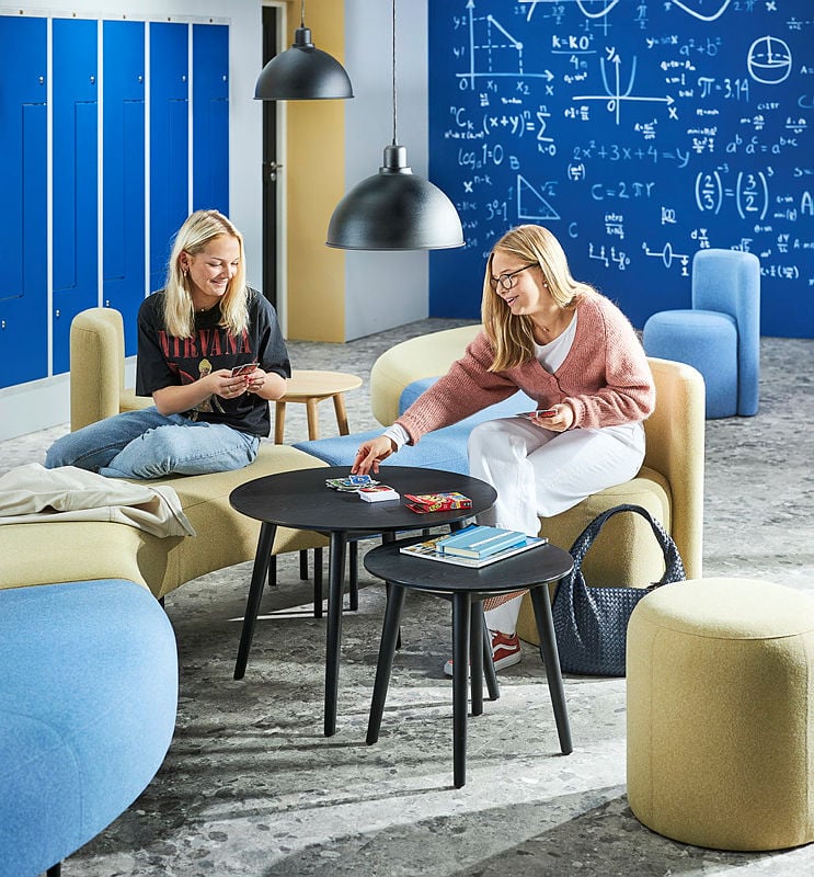 A vibrant and modern school breakout space where two students are engaged in a card game at a set of black round tables. One student, wearing a Nirvana T-shirt and jeans, is sitting on a curved yellow bench, smiling while holding her cards. The other student, in a pink knitted sweater and white trousers, is reaching for a card while seated on a matching yellow seat. The room features contemporary furniture, including soft seating in shades of blue and yellow, black pendant lights, and a blue chalkboard wall filled with mathematical equations and scientific diagrams. Blue lockers are lined up against the wall, adding to the academic setting. The area is designed for relaxation and informal learning, encouraging social interaction and creativity.