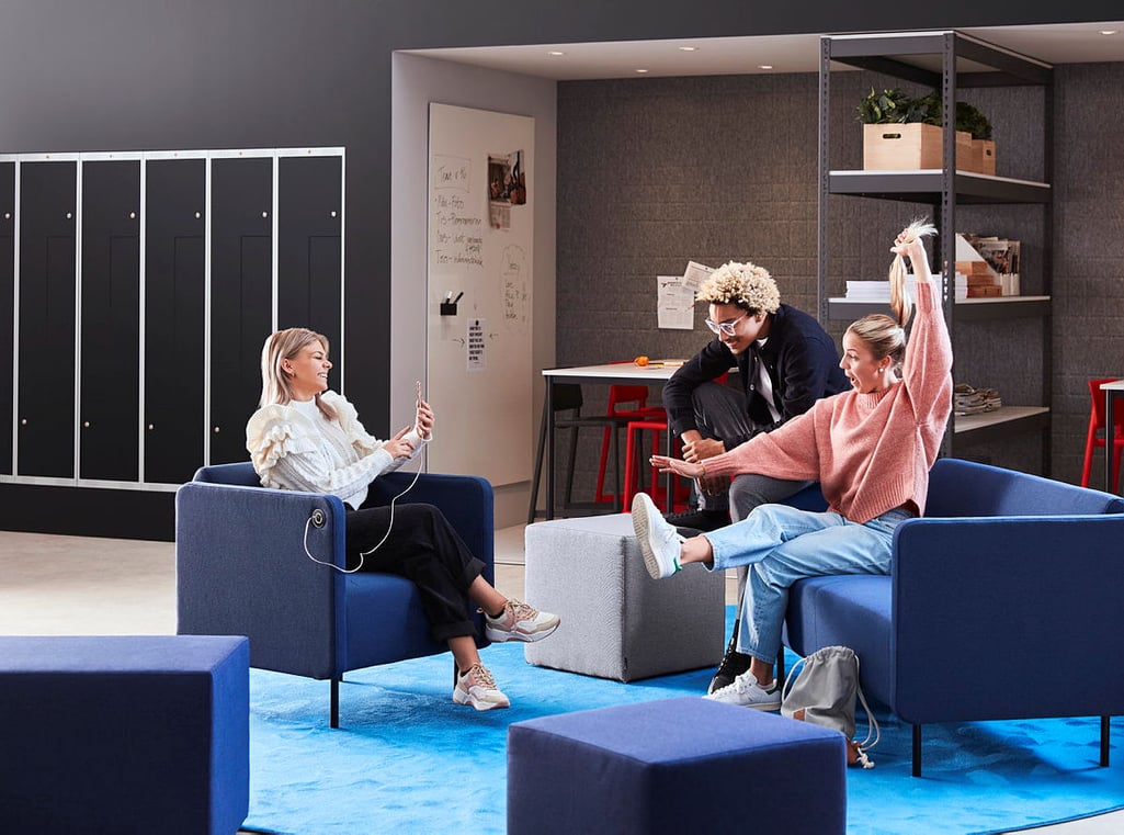 A lively and modern school breakout room where three students are socializing in a relaxed setting. The space is furnished with comfortable blue armchairs, grey ottomans, and a vibrant blue carpet. One student, wearing a pink sweater and jeans, playfully stretches her arms while chatting with another student in a white ruffled sweater, who is seated and holding a phone with earphones. A third student, with curly blonde hair and glasses, is leaning in from a nearby stool, engaging in the conversation. The background features black lockers, a collaborative workspace with red stools, a whiteboard with notes, and industrial-style shelves stocked with books, plants, and storage boxes. The environment is open, inviting, and designed for students to unwind, collaborate, or work on projects in a casual atmosphere.