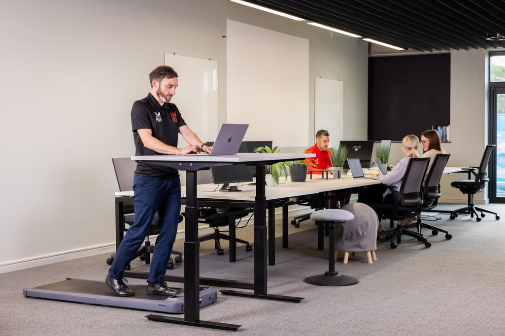 Man using a treadmill desk in a modern office, with colleagues working at desks in the background.