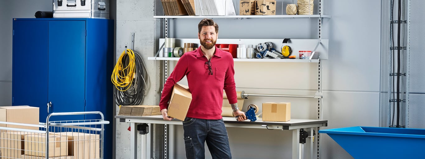 A man standing in a workspace, smiling while holding a cardboard box. The background includes a blue cabinet, shelves with various tools and supplies and a table. There are also several stacks of cardboard boxes and a blue container in the scene.