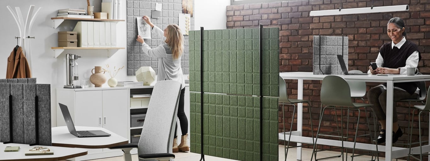 modern, minimalist office space. Two women are working; one is organizing documents on a pinboard, the other is seated at a high table using her phone. The office features contemporary furniture, including high stools, a shared work table, and room dividers that create a sense of both privacy and openness. The color palette is neutral and calming, primarily consisting of whites, grays, and muted greens. The overall atmosphere appears calm, collaborative, and professional.