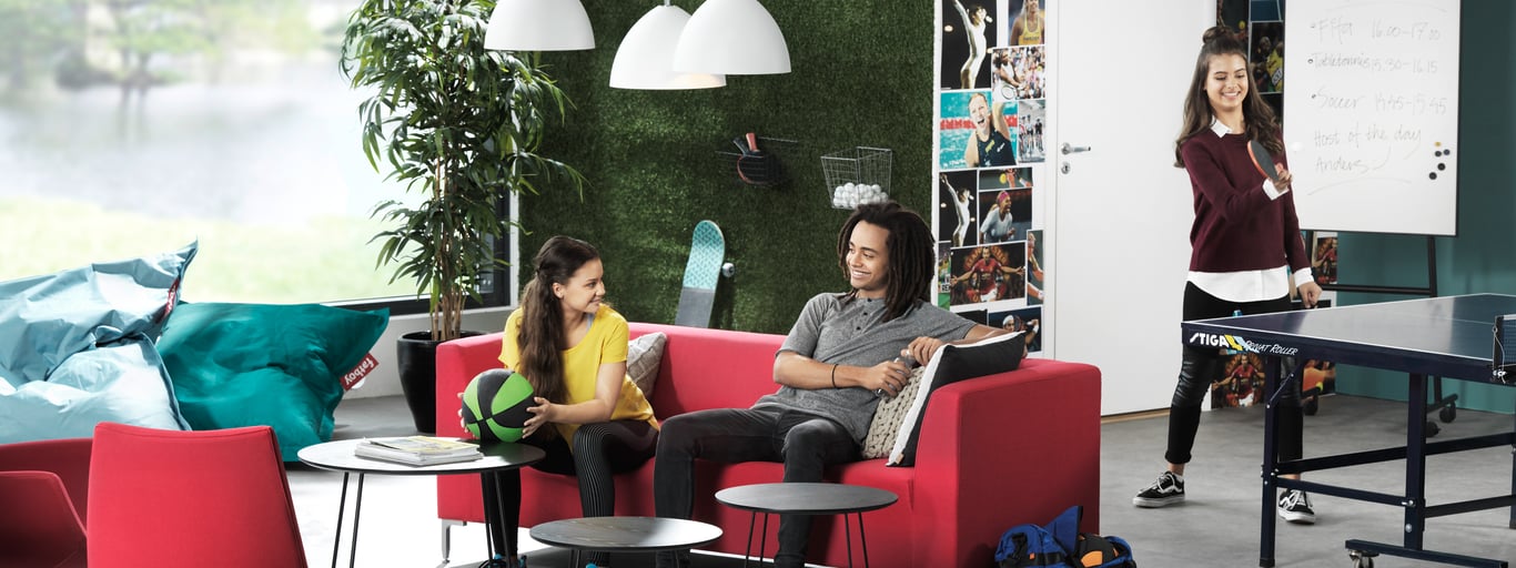 A modern, collaborative workspace featuring a casual seating area with red sofas and black tables. Two people are sitting on the sofas engaged in conversation, while another person stands in the background near a flip chart, actively presenting. The room is designed with a vibrant and creative atmosphere, including a green plant wall, pendant lights, and a large window allowing natural light to enter. The setting suggests an informal yet productive environment, ideal for teamwork and brainstorming sessions.