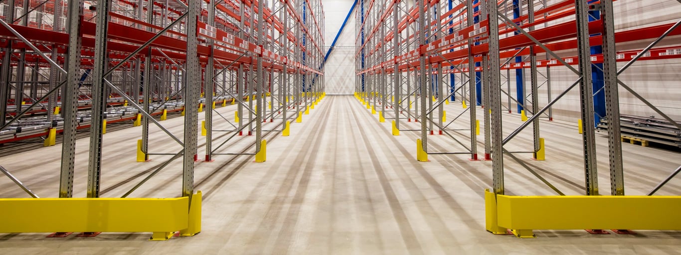 Full indoor shot of a large warehouse or cold storage facility. Metal racking systems, painted red, are arranged in tightly spaced, double rows. The racking system spans the entire image width, creating a long, narrow aisle down the center. Yellow plastic or rubber bumpers/protectors are attached to the concrete floor along the bases of all the metal racking columns. The image's focus is on the structural design and organization of the storage space, with a clear view of the metal framework and the defined aisle in the center. Industrial efficiency and robust storage capacity with AJ Products racking shelves