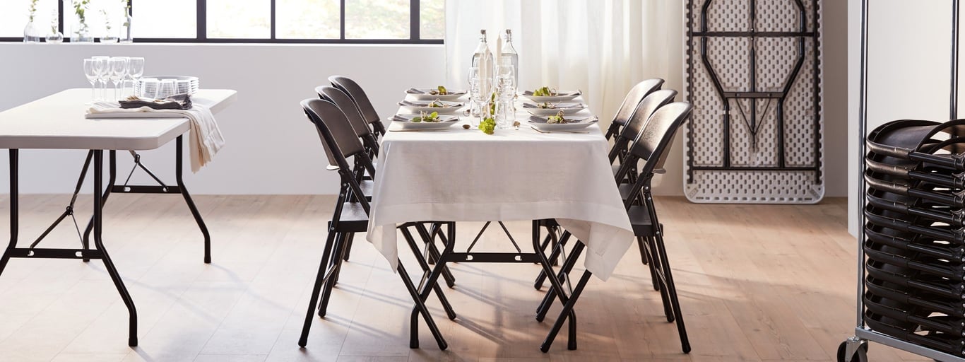 Minimalist, modern setting with a rectangular folding table draped with a white tablecloth. Eight black folding chairs are arranged around the table, which is set for a dinner party with plates, glasses, and simple centerpieces