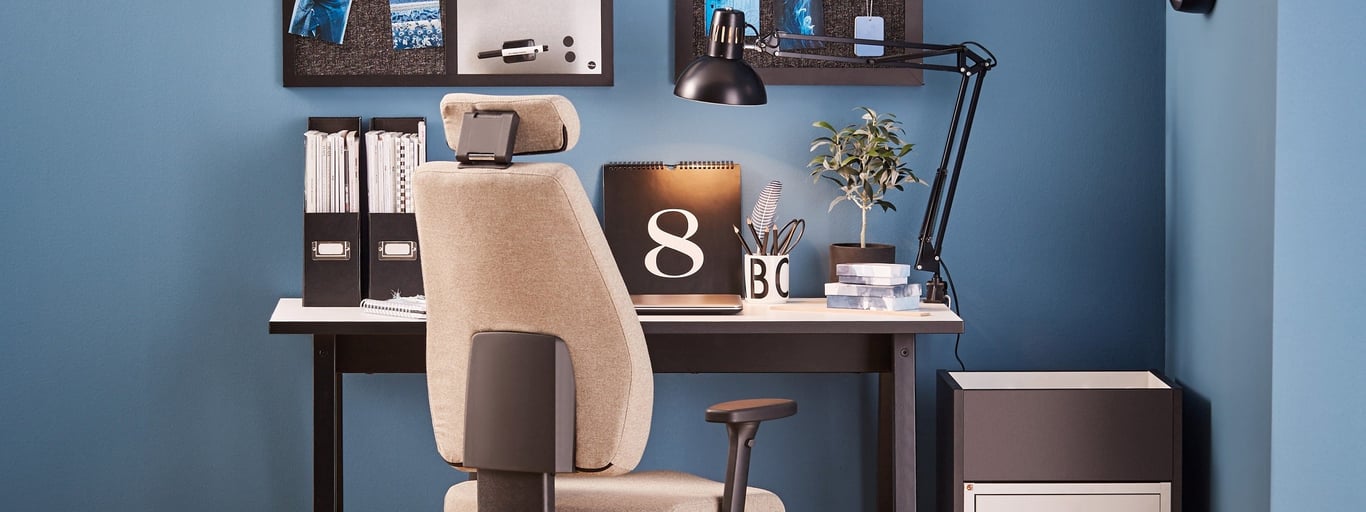 A stylish home office setup featuring a beige ergonomic chair with a headrest, positioned in front of a black desk. The desk is adorned with neatly organised files, a desk lamp, and decorative elements, including a plant and a calendar. The backdrop includes a vibrant blue wall and a pinboard displaying notes and photos, creating a modern and productive workspace.