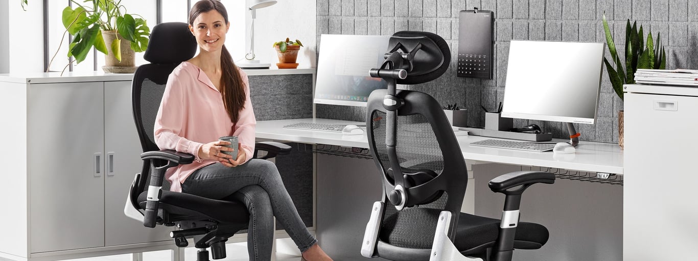 A modern office setup featuring a woman seated on a black ergonomic mesh chair with an adjustable headrest, holding a cup and smiling. The workspace includes a clean white desk with dual monitors, acoustic grey panels, and potted plants, creating a stylish, comfortable, and productive environment.