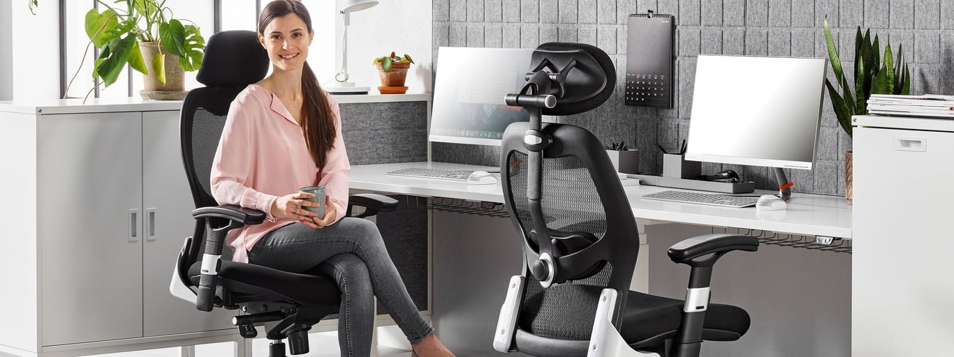 A modern office setup featuring a woman seated on a black ergonomic mesh chair with an adjustable headrest, holding a cup and smiling. The workspace includes a clean white desk with dual monitors, acoustic grey panels, and potted plants, creating a stylish, comfortable, and productive environment.