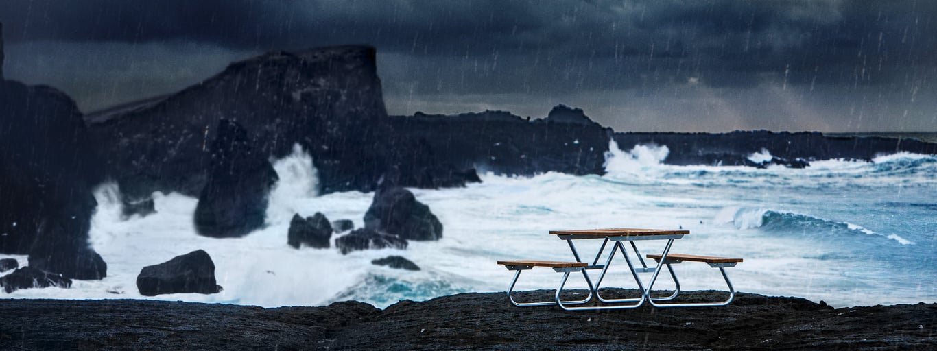 outdoor picnic table in a rainstorm. In the background, large waves crash against rocky outcroppings jutting out into the ocean. The sky is dark and overcast.