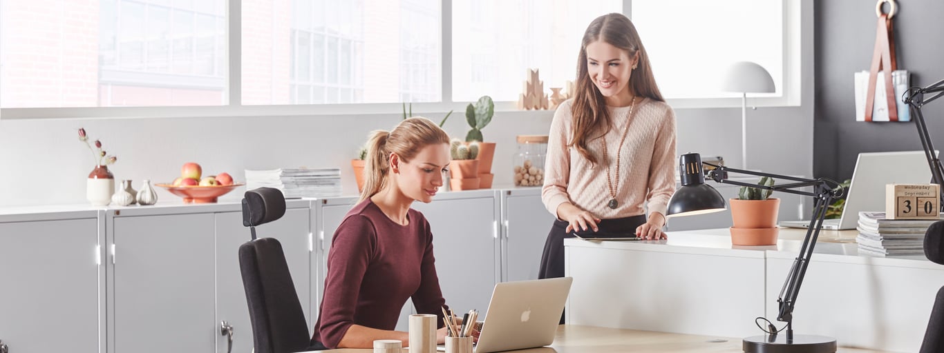 Two young women in a modern, bright office setting. One woman is seated at a desk working on a laptop, while the other stands beside her offering assistance