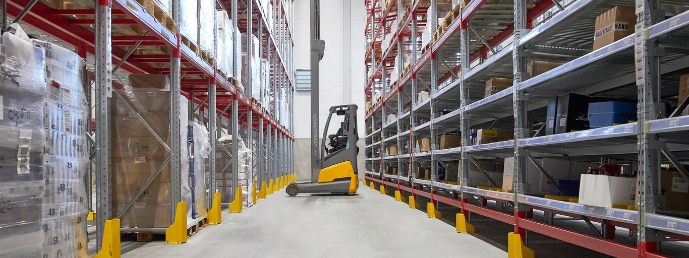 A warehouse aisle with tall, narrow shelves filled with boxes. A forklift is positioned in the center of the aisle moving goods within the facility.