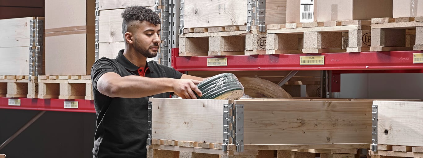 a young man wearing a black polo, carefully placing an item, into a wooden crate. The crate sits on a pallet within a larger warehouse setting filled with other stacked crates and pallets on shelving units.