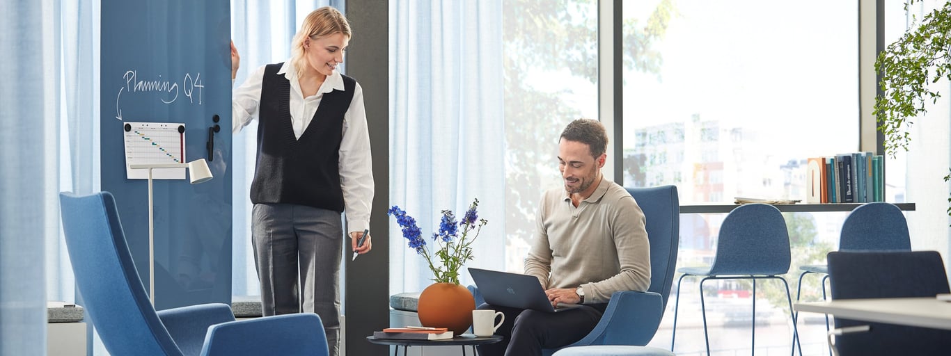 a woman standing beside a glass board in a meeting with a man that is looking at his laptop. They are engaged in a collaboration related to their work