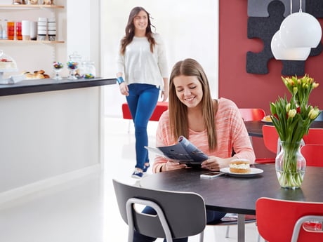 "A bright and welcoming breakroom where a young woman with long brown hair, wearing a pink sweater, is seated at a table reading a magazine with a smile. In front of her is a plate with a snack and a vase filled with fresh tulips. In the background, another person in blue jeans and a white top is walking towards the counter, which is stocked with various refreshments. The room features red chairs and a modern design, creating a comfortable and inviting atmosphere for relaxation and casual conversations.