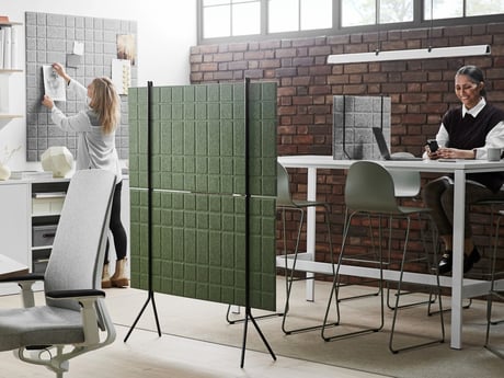 modern, minimalist office space. Two women are working; one is organizing documents on a pinboard, the other is seated at a high table using her phone. The office features contemporary furniture, including high stools, a shared work table, and room dividers that create a sense of both privacy and openness. The color palette is neutral and calming, primarily consisting of whites, grays, and muted greens. The overall atmosphere appears calm, collaborative, and professional.