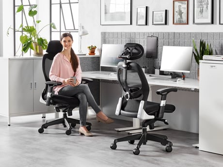 A modern office setup featuring a woman seated on a black ergonomic mesh chair with an adjustable headrest, holding a cup and smiling. The workspace includes a clean white desk with dual monitors, acoustic grey panels, and potted plants, creating a stylish, comfortable, and productive environment.