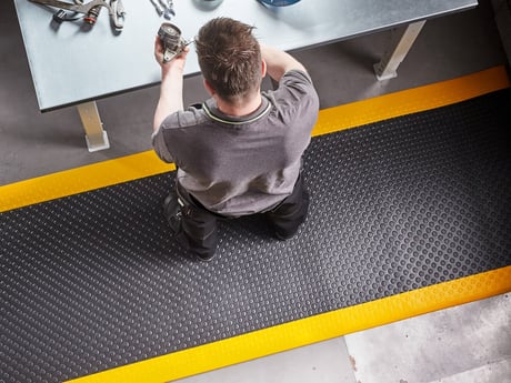 A person standing thoughtfully in a workspace, with a textured, dark rubber mat beneath their feet, bordered by bright yellow lines