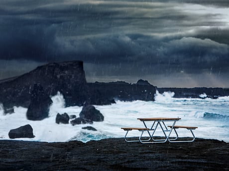 outdoor picnic table in a rainstorm. In the background, large waves crash against rocky outcroppings jutting out into the ocean. The sky is dark and overcast.