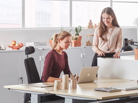 Two young women in a modern, bright office setting. One woman is seated at a desk working on a laptop, while the other stands beside her offering assistance