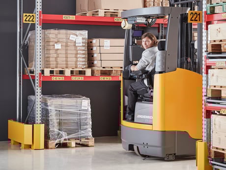 A warehouse worker operating a yellow forklift in a warehouse. The worker is seated in the forklift, which is positioned amidst high shelving units stocked with boxes and pallets of goods. A workday scene in a typical warehouse or distribution center.