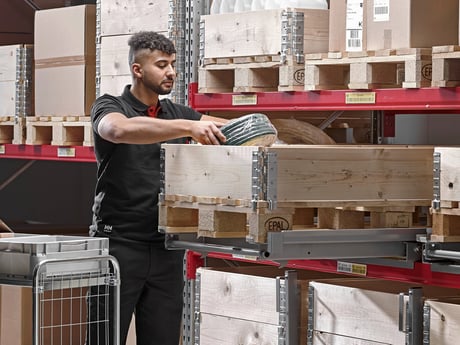 a young man wearing a black polo, carefully placing an item, into a wooden crate. The crate sits on a pallet within a larger warehouse setting filled with other stacked crates and pallets on shelving units. 