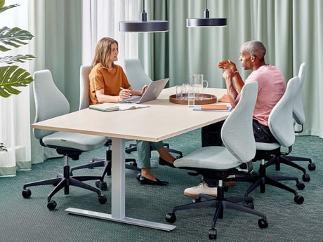 Divi kolēģi sarunājas pie konfeenču galda modernā gaiši zaļā birojā