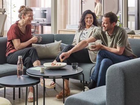 Three people sitting on a gray sectional sofa engaged in conversation and enjoying some refreshments.