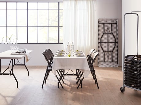 Minimalist, modern setting with a rectangular folding table draped with a white tablecloth. Eight black folding chairs are arranged around the table, which is set for a dinner party with plates, glasses, and simple centerpieces