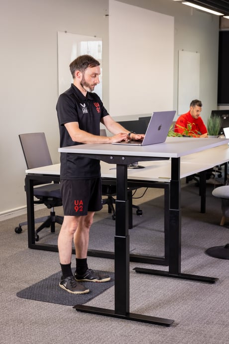 Man working at a standing desk with a laptop, wearing UA92-branded athletic gear.