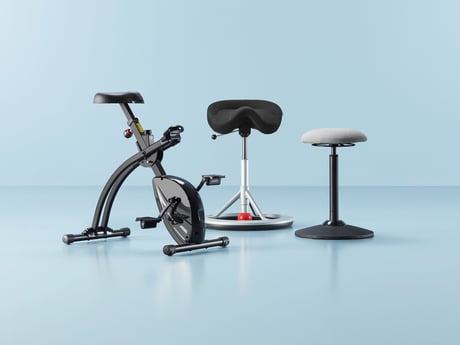 A set of three ergonomic and active seating solutions displayed against a light blue background. On the left, a sleek black office desk bike with a compact design and adjustable seat is positioned for workplace fitness. In the center, a black saddle-style stool with a white base and caster wheels offers dynamic seating for improved posture. On the right, a height-adjustable wobble stool with a round cushioned seat and a slightly curved base promotes active sitting. The collection highlights modern office furniture designed for movement, comfort, and improved ergonomics in the workplace.